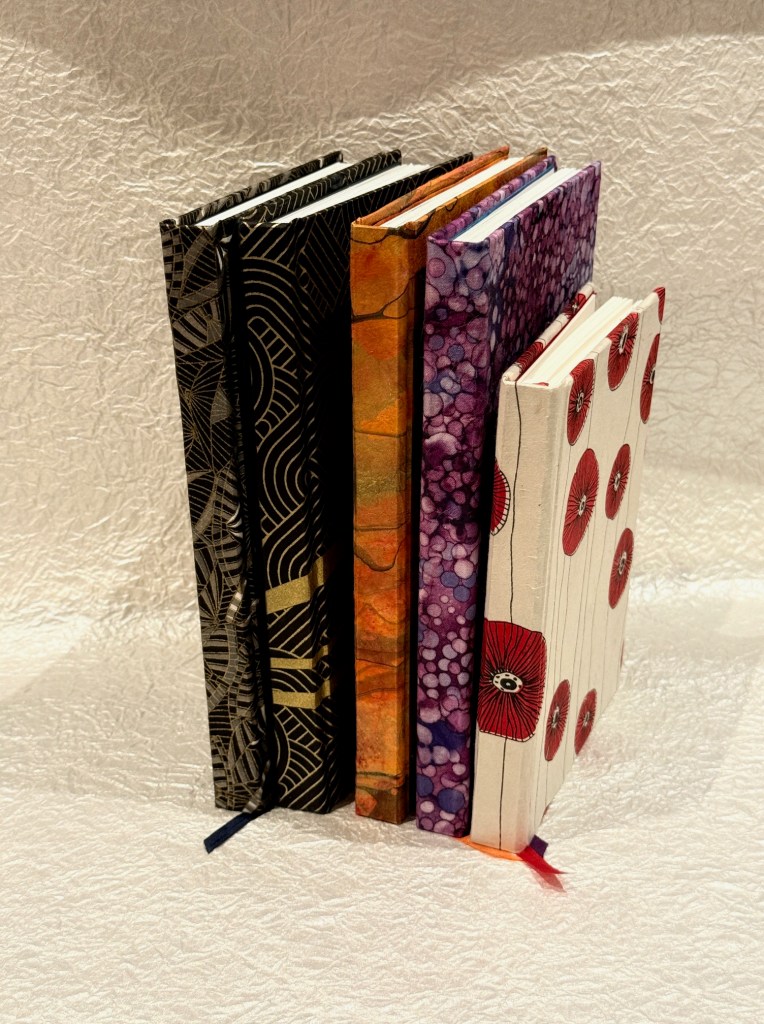 A collection of five decorative notebooks stacked side by side, showcasing various colorful covers and designs against a textured background.
