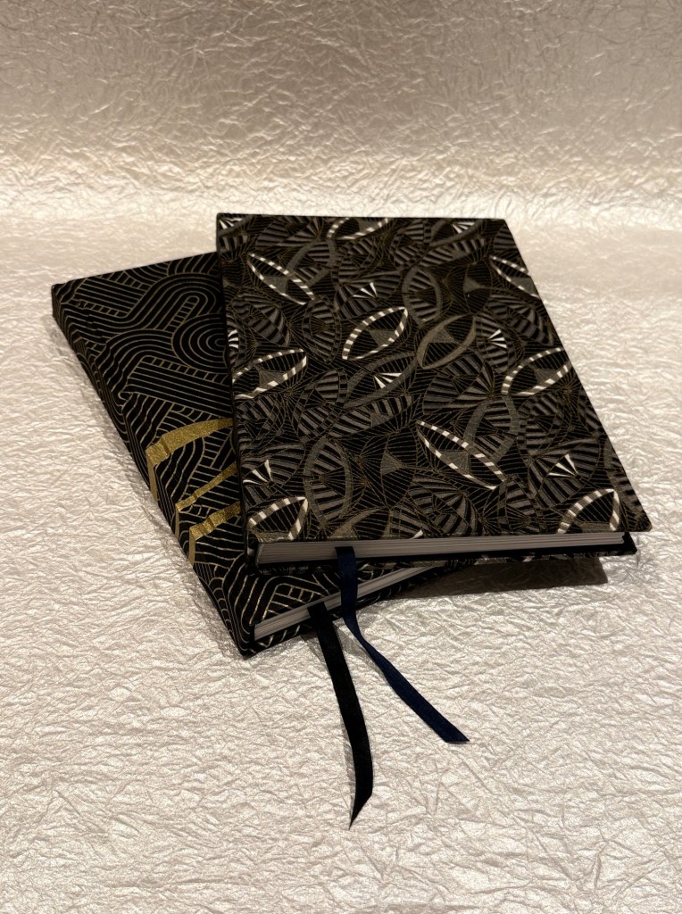 Two decorative notebooks with patterned covers, one featuring a leaf design in shades of black and gold, and the other with a geometric design, placed on a textured silver background.